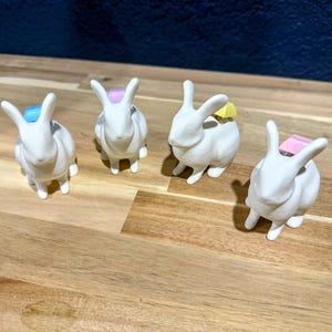 May include: Four white ceramic bunny figurines with different colored paper clips on their heads. The bunny figurines are arranged on a wooden surface.