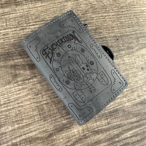 Pode incluir: Carteira de couro cinza com a palavra "ENCHIRIDION" e um desenho de caveira e espada. A carteira tem uma alça preta e é retangular. O desenho é gravado no couro.
