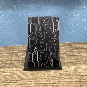 Könnte beinhalten: Ein schwarzer und weißer Kartenhalter aus Metall mit einem Leiterplatten-Design. Der Kartenhalter wird auf einem schwarzen Ständer präsentiert. Der Hintergrund ist eine hellblaue Wand und eine Holzoberfläche.