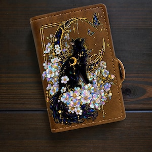 May include: A brown leather-like notebook cover featuring a printed illustration of a black cat sitting amidst iridescent white flowers and a golden crescent moon. Two small butterflies are depicted above the cat, with gold-toned accents.