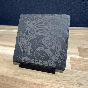 May include: A black slate coaster with a gray engraved design of a dragon. The coaster is on a wooden surface and has a black stand.