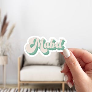 May include: A sticker with the name "Mabel" written in a retro, cursive font. The letters are outlined in green and filled with a light beige color.