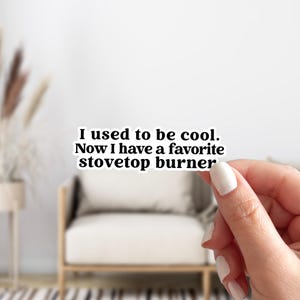 May include: A white sticker with black text that reads, "I used to be cool. Now I have a favorite stovetop burner." The sticker is held by a person with a blurred background of a chair and plant.