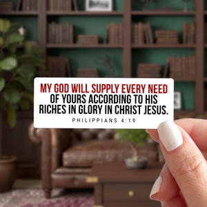 May include: A white rectangular sticker with the text "MY GOD WILL SUPPLY EVERY NEED OF YOURS ACCORDING TO HIS RICHES IN GLORY IN CHRIST JESUS. PHILIPPIANS 4:19" in black and red font. The sticker is held by a person with a blurred background.