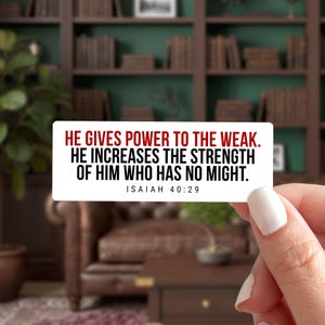 May include: A rectangular white sticker with the text "HE GIVES POWER TO THE WEAK. HE INCREASES THE STRENGTH OF HIM WHO HAS NO MIGHT. ISAIAH 40:29" in red and black. The sticker is held by a person with a blurred background of a room with bookshelves.