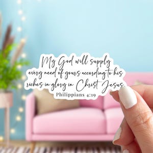 May include: A white sticker with the text "My God will supply every need of yours according to his riches in glory in Christ Jesus. Philippians 4:19" in black script. The sticker is held in front of a pink sofa and a light blue background.