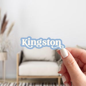 May include: A light blue sticker with the name "Kingston" in white script. The sticker has a light blue border and is held by a person with white nail polish. The background includes a white chair and neutral decor.