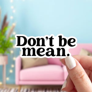 May include: A white sticker with the words "Don't be mean" in bold, black letters. The sticker is held by a person's hand with a pink couch and blue background.