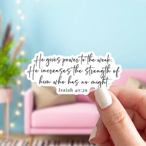 May include: A white sticker with black script. The text reads: "He gives power to the weak. He increases the strength of him who has no might. Isaiah 40:29." The sticker is held by a person with a pink couch in the background.