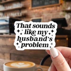 May include: A white sticker with black text that reads "That sounds like my husband's problem." The sticker has a black border with star accents. The background is a blurred image of a coffee shop.