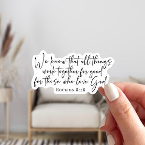 May include: A white sticker with black script that reads "We know that all things work together for good for those who love God" from Romans 8:28. The sticker is held by a person with a blurred background.