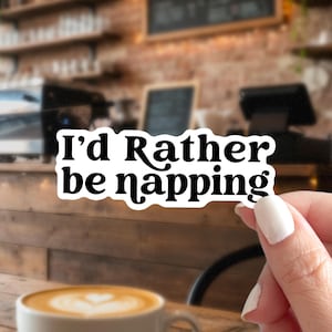 May include: A die-cut sticker with a white background and black text that reads I'd Rather be napping in a bold, rounded, retro-style font. The sticker features a clean white border around the lettering, creating a distinct contrast for the text.