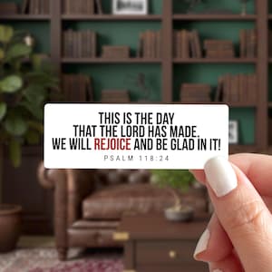 May include: A white rectangular sticker with the text "THIS IS THE DAY THAT THE LORD HAS MADE. WE WILL REJOICE AND BE GLAD IN IT!" and "PSALM 118:24" in black and red. The sticker is held in front of a blurred background.