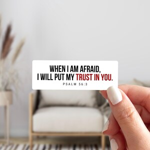 May include: A rectangular sticker with the text "WHEN I AM AFRAID, I WILL PUT MY TRUST IN YOU. PSALM 56:3" in black and red. The sticker is held by a hand with a white painted nail.