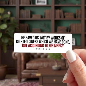 May include: A white rectangular sticker with the text "HE SAVED US, NOT BY WORKS OF RIGHTEOUSNESS WHICH WE HAVE DONE, BUT ACCORDING TO HIS MERCY TITUS 3:5" in black and red. The sticker is held by a person with a blurred background of a room with bookshelves.