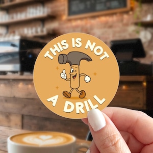 May include: A round sticker with a cartoon hammer character giving a thumbs up. The sticker is orange with the text "THIS IS NOT A DRILL" in a white, playful font. The hammer has a brown handle, gray head, and is winking.