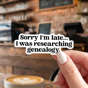 May include: A white sticker with black text that reads, "Sorry I'm late... I was researching genealogy." The sticker is held by a person's hand in front of a blurred coffee shop background.