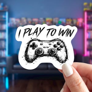 May include: A white sticker featuring a black and white illustration of a video game controller. The text "I PLAY TO WIN" is written above the controller in a bold, stylized font. The sticker has a hand-drawn, sketched appearance.