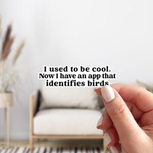 May include: A white sticker with black text that reads, "I used to be cool. Now I have an app that identifies birds." The sticker is held by a person with manicured nails. The background is a blurred interior scene.