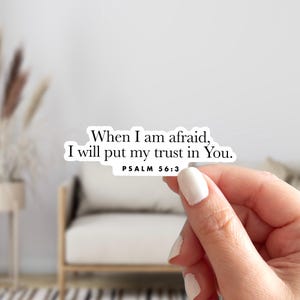 May include: A white sticker with the text "When I am afraid, I will put my trust in You. PSALM 56:3" in black. The sticker is held by a hand with white nail polish. The background is a blurred interior scene.