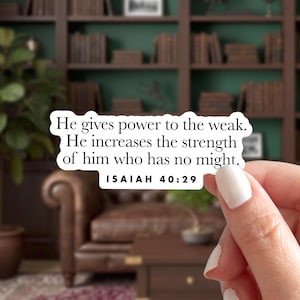 May include: A white sticker with the text "He gives power to the weak. He increases the strength of him who has no might. ISAIAH 40:29." The sticker is held by a person with white nail polish. The background is a blurred interior with bookshelves.