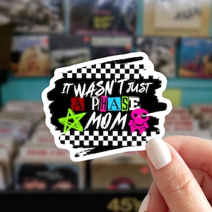 May include: A sticker with the text "IT WASN'T JUST A PHASE MOM" in various colors. The sticker has a black background with a white border and a checkered pattern. A neon green star and a pink skull and crossbones are also featured.