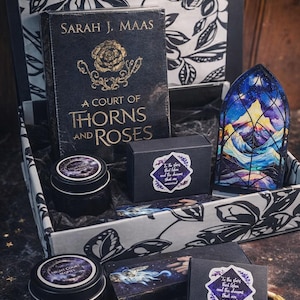 May include: A curated gift box featuring a book titled "A Court of Thorns and Roses" by Sarah J. Maas, candles, and other themed items. The box has a black and white floral design, with additional items in black and gold.