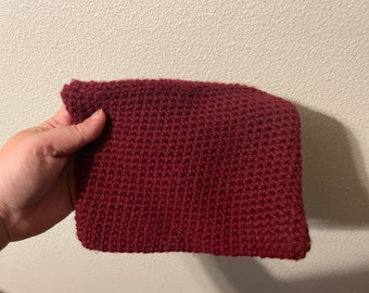 Puede incluir: Un cuadrado tejido a mano de color burdeos. La textura del hilo es visible, con cada puntada creando un patrón en relieve. El artículo se sostiene en una mano, mostrando el tamaño y la forma de la pieza tejida.