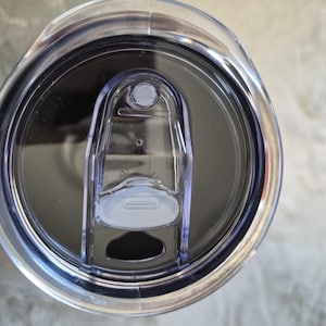 May include: A clear plastic tumbler lid with a sliding closure. The lid has a dark grey interior and a clear plastic slider to open and close the drinking hole. The lid is designed to fit a standard tumbler.