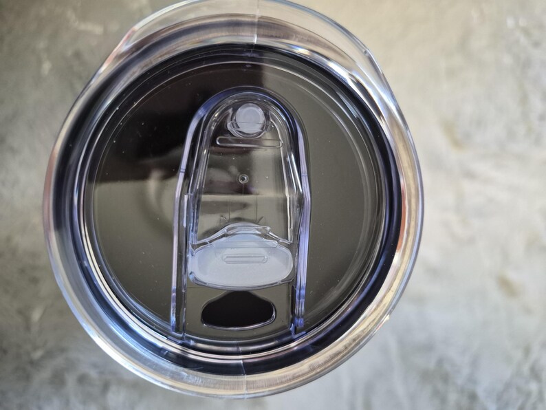 May include: A clear plastic tumbler lid with a sliding closure. The lid has a dark grey interior and a clear plastic slider to open and close the drinking hole. The lid is designed to fit a standard tumbler.