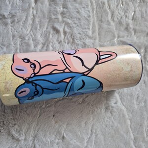 May include: A cylindrical tumbler with a cartoon design. The design features a pink and blue cartoon character lying down on a sandy background. The tumbler is likely made of stainless steel and is suitable for cold drinks.