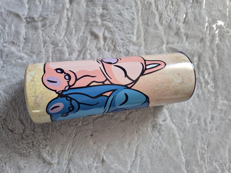 May include: A cylindrical tumbler with a cartoon design. The design features a pink and blue cartoon character lying down on a sandy background. The tumbler is likely made of stainless steel and is suitable for cold drinks.