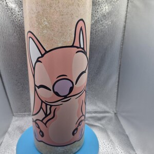 May include: A stainless steel tumbler with a pink cartoon character design. The character has large ears and a purple nose. The tumbler has a light brown speckled background and sits on a blue base. The character is smiling with its eyes closed.