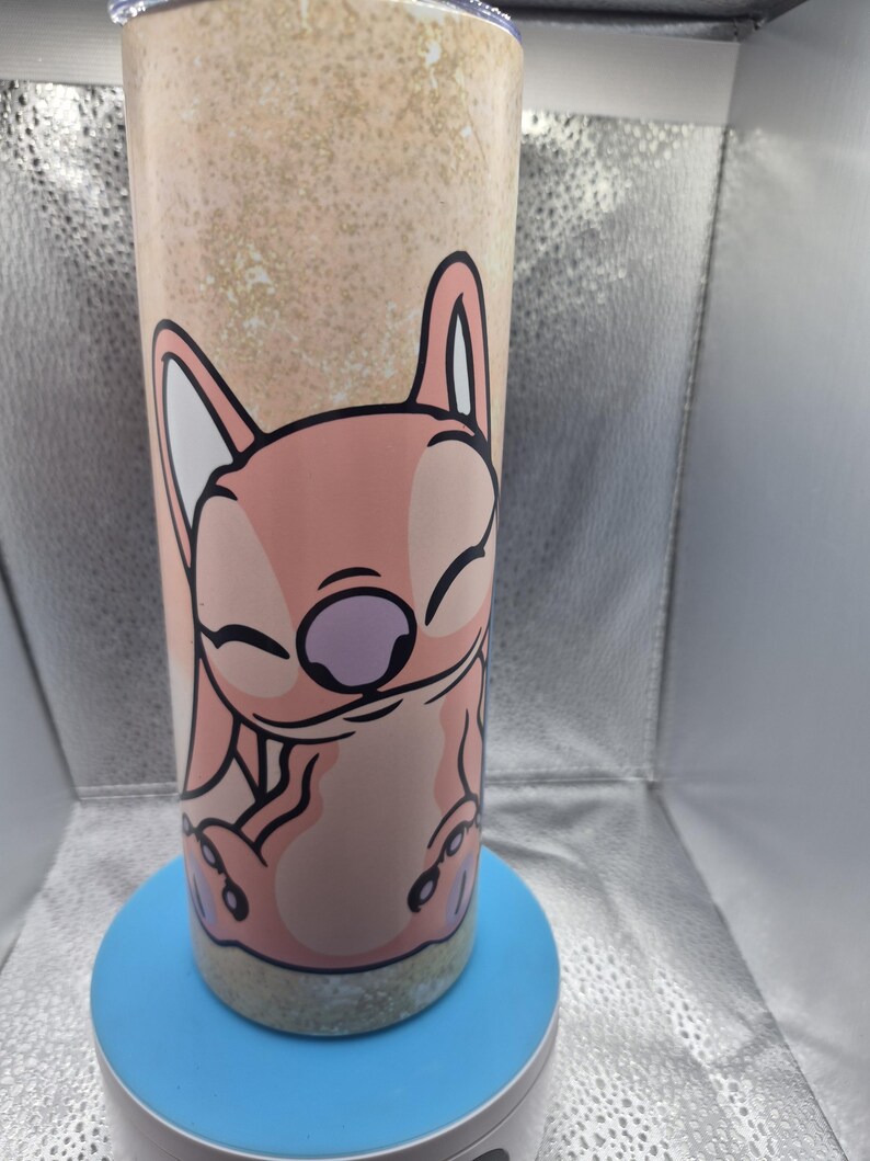 May include: A stainless steel tumbler with a pink cartoon character design. The character has large ears and a purple nose. The tumbler has a light brown speckled background and sits on a blue base. The character is smiling with its eyes closed.