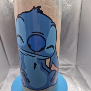May include: A blue tumbler with a cartoon Stitch design. The tumbler has a light blue base and a background with a gradient of peach, gold, and light blue. The cylindrical tumbler features the character Stitch with a smiling expression.