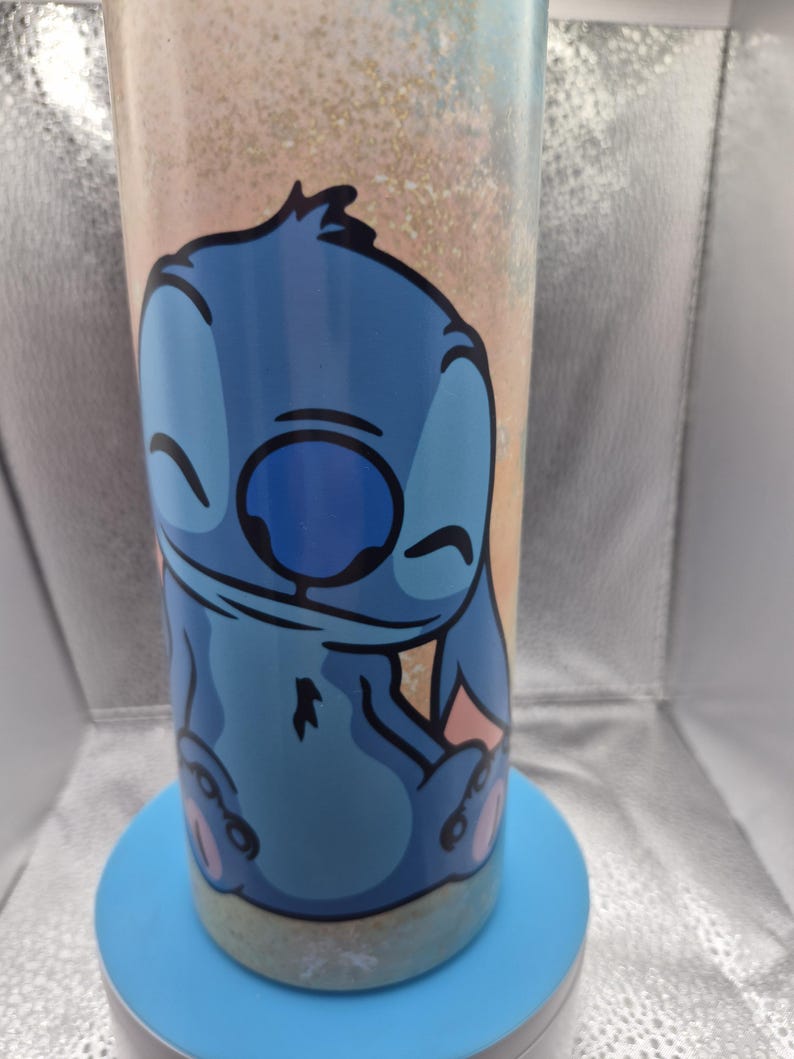 May include: A blue tumbler with a cartoon Stitch design. The tumbler has a light blue base and a background with a gradient of peach, gold, and light blue. The cylindrical tumbler features the character Stitch with a smiling expression.