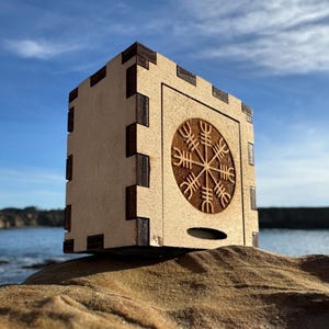 Könnte beinhalten: Eine hellfarbene Holzkiste mit dunkelbraunen Akzenten. Die Kiste hat ein geschnitztes kreisförmiges Design auf der Vorderseite und ist mit ineinandergreifenden Verbindungen konstruiert. Die Kiste steht auf einem Felsen mit blauem Himmel und Ozean im Hintergrund.