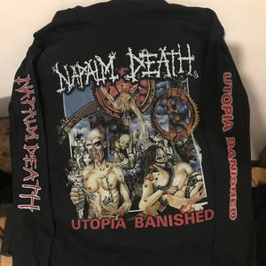 Może przedstawiać: Czarna koszulka z długim rękawem z nazwą zespołu "NAPALM DEATH" w kolorze białym oraz napisem "Utopia Banished" pod grafiką postaci i maszyn. Na rękawach również nazwa zespołu i "Utopia Banished" w kolorze czerwonym.