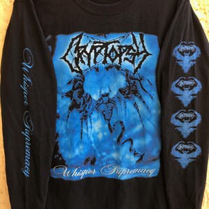 Pode incluir: Uma camiseta preta de manga comprida com um gráfico azul no peito com o texto Cryptopsy e Whisper Supremacy. A manga esquerda tem o texto Whisper Supremacy em letra cursiva azul, e a manga direita tem quatro ícones de logotipos alados azuis.