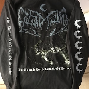 Può includere: Maglietta nera a maniche lunghe con un design grafico sulla schiena e sulle maniche. La schiena presenta un design stilizzato con la scritta "The Tenth Sub Level Of Suicide". Le maniche hanno disegni a mezzaluna.