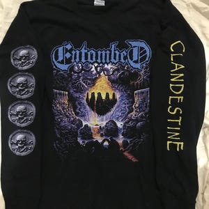 Puede incluir: Camiseta negra de manga larga con el nombre de la banda "Entombed" en azul y un gráfico de una escena de cueva con una cascada. La manga izquierda tiene cuatro emblemas de calavera y huesos cruzados, y la manga derecha dice "Clandestine" en amarillo.