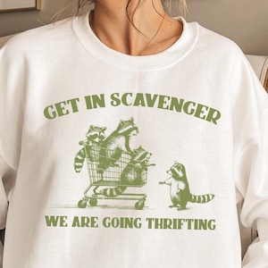 Op de afbeelding: Wit sweatshirt met een groene afbeelding van wasberen in een winkelwagen. De tekst "Get in Scavenger We are going thrifting" staat op het shirt gedrukt.