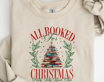 May include: A beige sweatshirt with a Christmas-themed design. The design features a stack of books shaped like a Christmas tree with the text "All Booked for Christmas Holly Jolly Book Club".