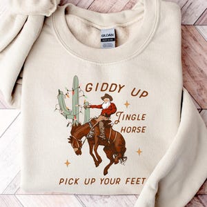May include: A beige sweatshirt with a graphic of a cowboy riding a bucking horse, a cactus with Christmas lights, and the text "Giddy Up, Jingle Horse, Pick Up Your Feet".