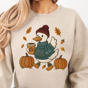 May include: Beige sweatshirt with an embroidered duck wearing a hat and scarf, holding a coffee cup. The design includes pumpkins, leaves, and acorns, evoking an autumnal theme. The sweatshirt is a comfortable, casual style.