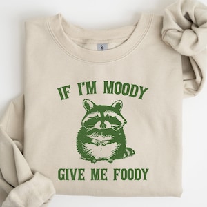 May include: A beige sweatshirt with a green graphic of a raccoon and the text "If I'm Moody Give Me Foody".