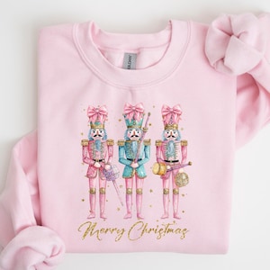 Könnte beinhalten: Rosafarbener Pullover mit einem Motiv von drei Nussknackern in Rosa, Blau und Gold, die Musikinstrumente halten. Der Text "Merry Christmas" ist in Gold unter den Nussknackern gedruckt.