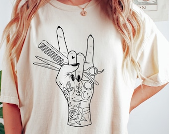 Könnte beinhalten: Ein weißes T-Shirt mit einer schwarzen Grafik einer Hand, die einen Kamm und eine Schere hält. Die Hand macht eine Rock'n'Roll-Handgeste. Die Hand ist mit einem Schmetterling, einer Rose, einer Mondsichel und Sternen tätowiert.