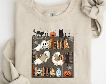 May include: Beige sweatshirt featuring a Halloween-themed graphic. The graphic depicts a bookshelf with ghosts reading books, black cats, pumpkins, candles, and spiderwebs. The sweatshirt has long sleeves and a crew neck.