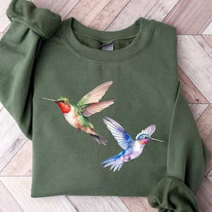 May include: A green sweatshirt with a watercolor design of two hummingbirds. One hummingbird is green, blue, and white, and the other is green, red, and white.